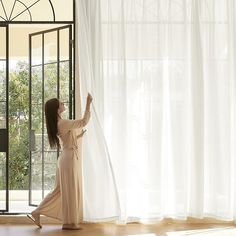 a woman standing in front of a window with her hand on the sheer curtain and looking out