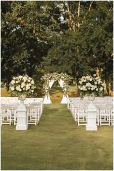 an outdoor ceremony setup with white chairs and flowers on the aisle, surrounded by trees