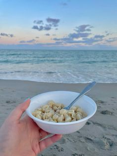 a hand holding a bowl of cereal on the beach at sunset with ocean in background