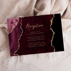 a red and black wedding card on top of a white blanket with gold foil lettering