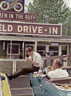 two men sitting in an old car at the drive - in, talking on their cell phones