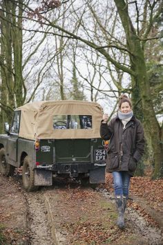 a woman standing next to an army truck in the woods with her hand up and smiling at the camera