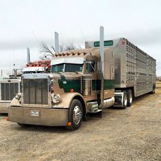 two large semi trucks parked next to each other on a dirt field with trees in the background