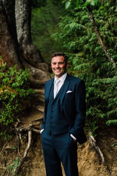 a man in a suit standing on a dirt path near some trees and bushes with his hands in his pockets