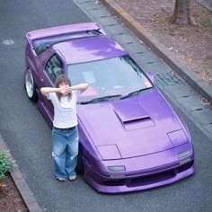 a woman standing next to a purple car on the side of a road with her arms behind her head