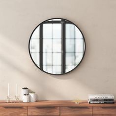 a round mirror hanging on the wall above a wooden dresser with books and vases