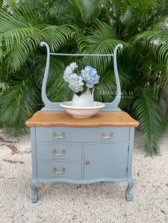 a blue dresser with flowers on top in front of some palm trees and plants behind it