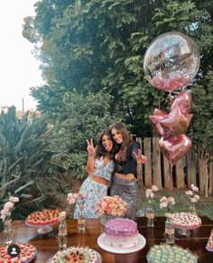 two women standing next to each other in front of a table with cakes and balloons