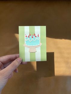 a hand holding up a card with a blue cake on it and red cherries