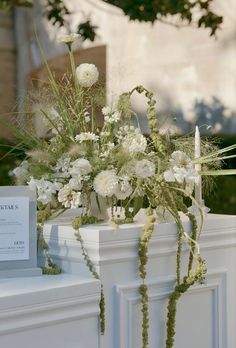 white flowers and greenery in a vase on a mantle with a sign that reads