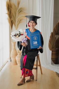 a woman sitting on a chair holding a plaque and flowers in her hand while wearing a graduation gown