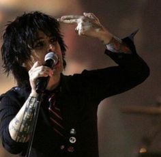 a man with black hair and piercings holding a microphone in his right hand while standing next to a drum set