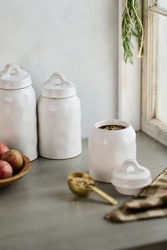 two white jars sitting on top of a counter next to apples and a bowl of fruit