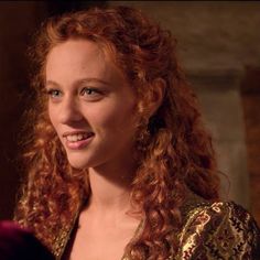 a woman with red curly hair smiling at the camera while wearing a gold dress and earrings