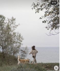 a woman is walking her dog on the grass by the water with trees in the background