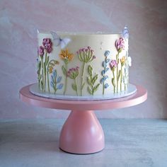 a white cake with colorful flowers and butterflies on it sitting on a pink stand in front of a wall