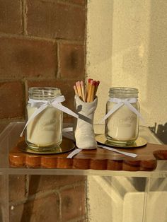 two mason jars with matchesticks tied to them sitting on a wooden shelf next to a brick wall