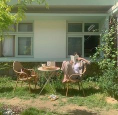 a woman sitting at a table reading a book outside in the grass next to a house