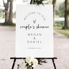 a welcome sign with white flowers on it and greenery in the foreground is an outdoor ceremony area