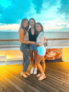 three girls are standing on a deck by the ocean with their arms around each other