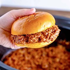 a hand holding a sloppy joe sandwich over a slow cooker filled with chili cheese