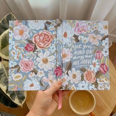 a person holding up a book with flowers on it and a cup of coffee in front of them