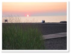 two people sitting on a bench at the beach watching the sun go down over the water