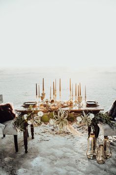 a table set up with candles and flowers on top of snow covered ground next to the ocean