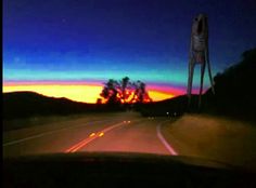 the sun is setting behind a skeleton statue on the side of the road with trees in the background