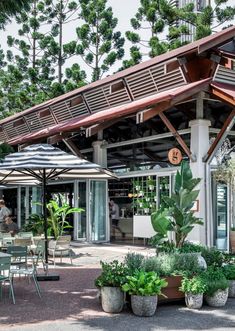 an outdoor cafe with tables and umbrellas on the patio, surrounded by plants and potted trees