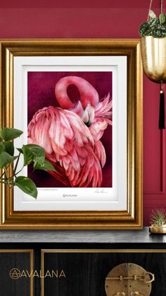 a pink flamingo in a gold frame sitting on top of a black cabinet next to a potted plant