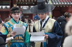 two women dressed in traditional chinese clothing and hats are looking at something on the paper