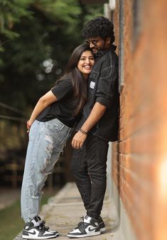 two people standing next to each other near a brick wall with trees in the background