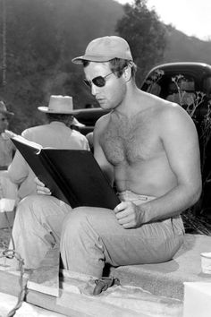 a shirtless man reading a book in front of a car while wearing sunglasses and a hat