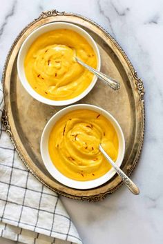 two white bowls filled with yellow hummus on top of a marble table next to silver spoons