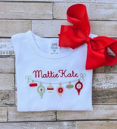 This adorable girl's shirt is perfect for Christmas and would be a super cute outfit for a Christmas card! The detailed design of the Christmas Ornaments, as well as your chosen name, is created by machine embroidery and uses high quality thread.  The white shirt is true to size and is made out of 100% combined cotton interlock fabric to keep your child completely comfortable. The shirt has naturally tapered shoulders and includes a ruffled hemline. The short sleeve shirts have puff sleeves.  Pl Christmas White Embroidered Tops, White Tops With Custom Embroidery For Christmas, Cute White Top For Festive Occasions, Embroidered Christmas Holiday Tops, Holiday Embroidered Tops As Gift, Holiday Cotton Top With Embroidery, Holiday Cotton Embroidered Tops, Red Embroidered Holiday Tops, White Christmas Tops With Embroidered Graphics