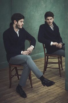 two men sitting on chairs in front of a wall, one with his legs crossed