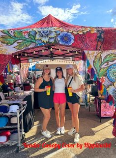 three women standing under a colorful tent at an outdoor market with drinks in their hands