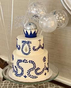 a three tiered cake with blue and white designs on it, surrounded by balloons