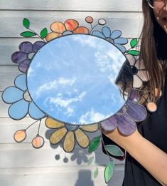 a woman holding up a mirror with flowers and clouds in the reflection, while looking at it