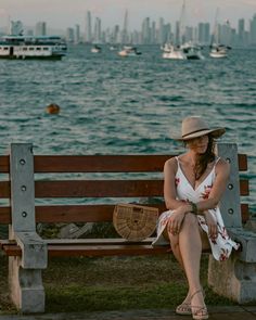 a woman in a hat sitting on a bench by the water with boats in the background