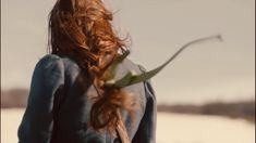 a woman with long red hair is walking in the snow holding a green plant behind her back