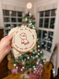 someone holding up a small embroidered christmas ornament in front of a decorated christmas tree