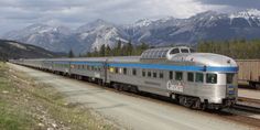 a silver and blue passenger train traveling down tracks next to snow covered mountain range in the distance