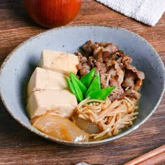 a bowl filled with noodles and meat on top of a wooden table next to an apple