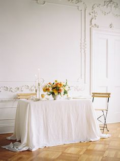 a table with flowers and candles on it in front of a white wall, next to a chair