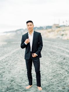 a man in a black suit and white shirt is standing on the beach with his hands in his pockets