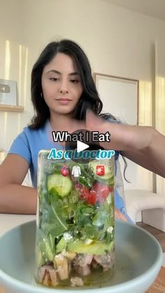 a woman sitting at a table in front of a glass jar filled with vegetables