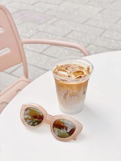 a cup of coffee and sunglasses sitting on a white table with a pink chair in the background