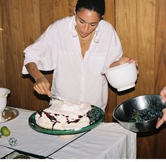 a woman in white shirt decorating cake with icing on table next to wooden wall